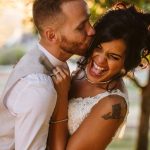 Bride laughs joyfully as the groom kisses her cheek, outdoors at sunset in wedding attire.
