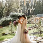Two brides in white gowns embrace and kiss in a sunlit garden, holding bouquets beneath a large chandelier.