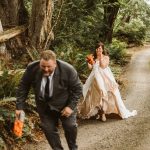 Bride in a white wedding dress chases a man in a suit along a forest path, both holding orange water guns.