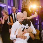 Bald man in a white shirt hugs a bride in a white wedding dress at a reception, with guests and purple lighting in the background.