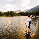 Couple embracing at the lake’s edge with forested hills and mountains in the background, sunset light.