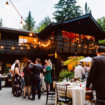 Snohomish Wedding Photographer GSquared Weddings A lively outdoor gathering at a house features guests mingling near tables with lights strung above. The backdrop is a large two-story building surrounded by trees. Everyone is dressed in formal attire, suggesting a celebratory event or party. Serving Seattle, Snohomish and Orlando