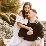 Couple sitting on sunlit rocky shoreline, woman in a white off-shoulder dress embraces a man in a black shirt.