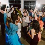 Wedding guests dance and raise their arms in a festive reception around the bride and groom with drinks in hand and tassel props, bright and lively.