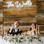 Bride and groom toasting at their head table with champagne, against a rustic wooden backdrop with greenery and candles.