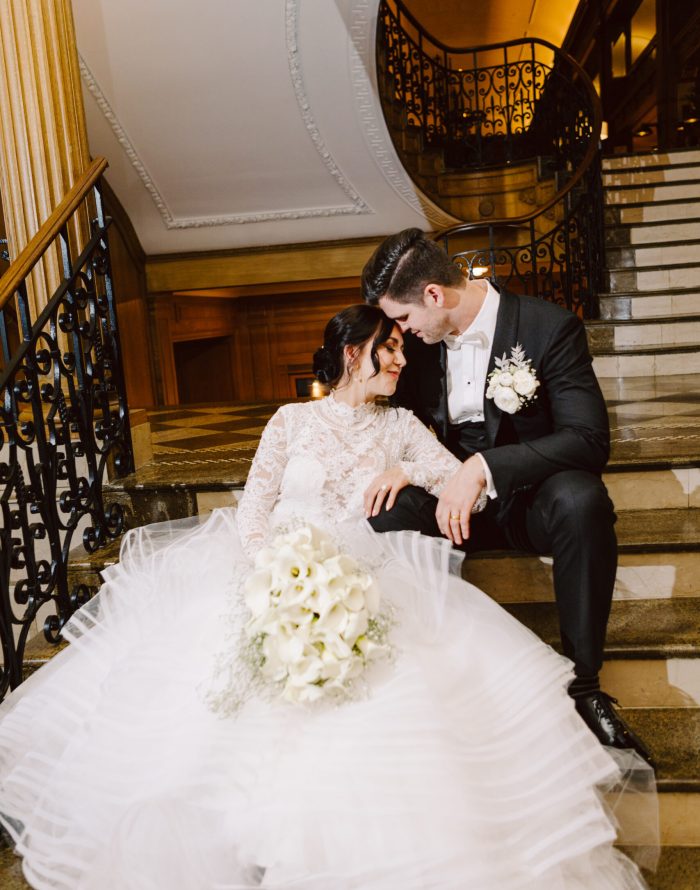 Fairmont Olympic Wedding Seattle Snohomish Wedding Photographer GSquared Weddings A bride in a white lace gown and a groom in a black tuxedo sit close together on elegant stairs, holding a bouquet of white flowers. Captured by Seattle Wedding Photography, they smile affectionately as they rest their heads together.