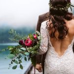 Bride in a lace back wedding dress holding a bouquet by a lake, facing away from the camera.