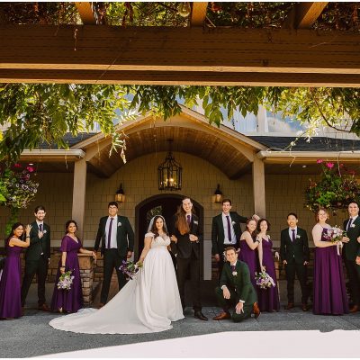 Group wedding party posed in matching purple dresses and green suits in front of a house entry.