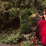 Couple in formal attire stands under a leafy apple tree; woman in red gown, man in dark suit.
