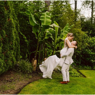 Snohomish Wedding Photographer GSquared Weddings In a lush Snohomish garden, a groom in a light suit lifts the bride in her white dress amid vibrant green foliage and banana plants. Their joy is palpable as she gazes at him lovingly while being held. Serving Seattle, Snohomish and Orlando