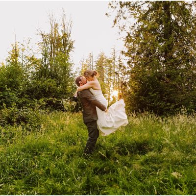 GSquared Weddings Photography A groom lifts and embraces his bride in a grassy, sunlit clearing surrounded by tall green trees in Snohomish County, both smiling and joyful on their wedding day. GSquared Weddings | 47.9129° N, 122.0982° W | Serving Snohomish, Seattle , Orlando & Beyond