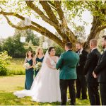 Bride and groom hold hands outdoors under a tree as bridesmaids in teal and groomsmen in dark suits stand nearby. A vintage chandelier hangs from the branches above.