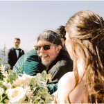 Smiling man wearing sunglasses hugs a woman outdoors at a wedding, with a bouquet in the foreground and guests in tuxedos in the background.